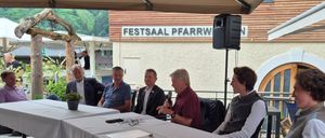 Fünf Männer sitzen um einen Tisch im Freien. Einer hält ein Mikrofon. Ein Schild darüber steht 'Festsaal Pfarrw'. Ein Lautsprecher ist auf einem Pfosten hinter ihnen montiert.