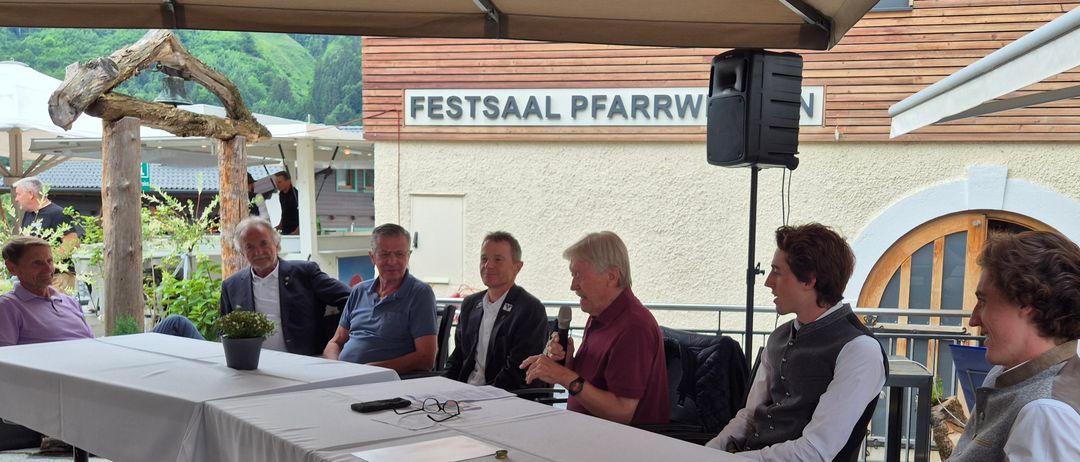 Fünf Männer sitzen um einen Tisch im Freien. Einer hält ein Mikrofon. Ein Schild darüber steht 'Festsaal Pfarrw'. Ein Lautsprecher ist auf einem Pfosten hinter ihnen montiert.