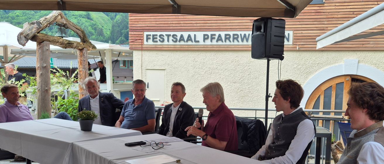 Fünf Männer sitzen um einen Tisch im Freien. Einer hält ein Mikrofon. Ein Schild darüber steht 'Festsaal Pfarrw'. Ein Lautsprecher ist auf einem Pfosten hinter ihnen montiert.