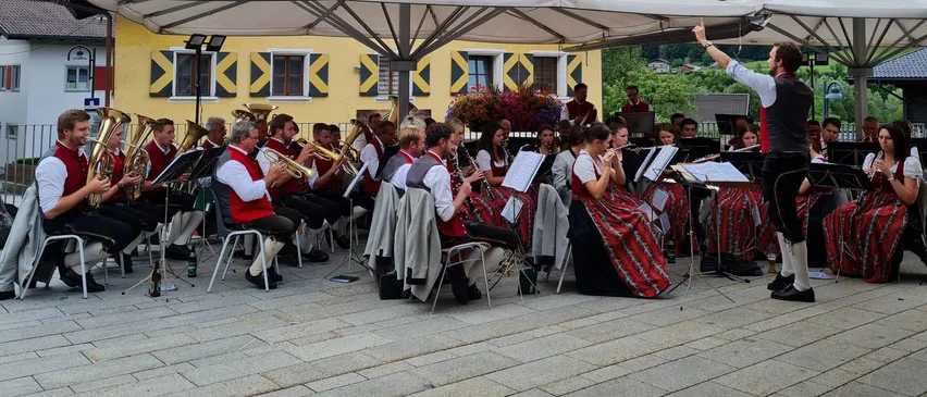 Ein Blasorchester spielt auf einem gepflasterten Platz, mit Musikern, die unter Sonnenschirmen vor einem gelben Gebäude sitzen. Ein Dirigent steht vorne und leitet das Ensemble.