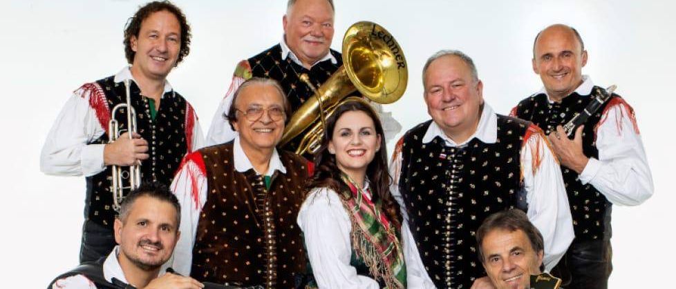 Ein Plakat zeigt eine Gruppe von Musikern, mit einer Frau und einem Mann in der Mitte, alle in traditioneller Kleidung, die verschiedene Instrumente halten. In der unteren linken Ecke befindet sich ein Regenschirm. Die Veranstaltung trägt den Titel "Dichtungwir." Sie ist für Freitag, den 31. Oktober 2025 geplant.