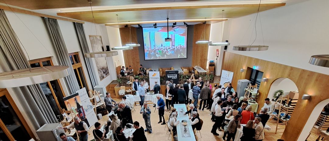 Eine Indoor-Veranstaltung mit einem großen Saal, einer Bühne und Menschen, die um Tische herum versammelt sind. Die Tische haben Weinflaschen, Gläser und Essen. Die Bühne hat eine Projektionsleinwand und ein Banner.