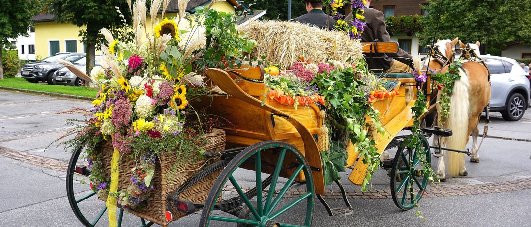 Ein Pferdewagen ist mit Blumen, Heu und Blättern geschmückt, zwei Personen sitzen darin. Der Wagen steht auf der Straße geparkt.