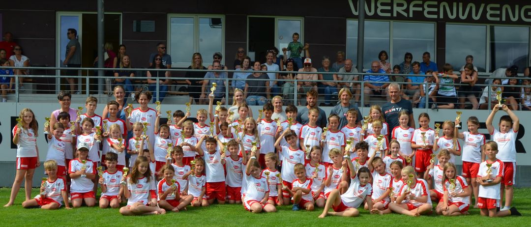 Eine Fußballmannschaft von Kindern posiert mit ihren Trophäen auf einem Feld für ein Foto. Dahinter sitzen Erwachsene auf Tribünen und schauen zu. Das Gebäude im Hintergrund hat 'Verfen' darauf.