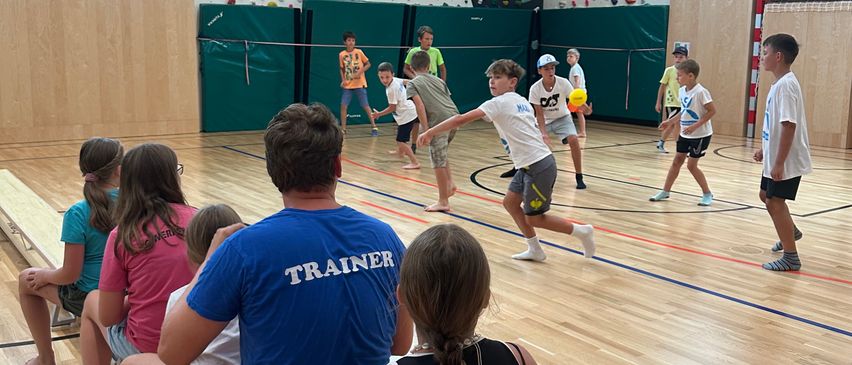 Eine Gruppe von Kindern und Erwachsenen spielt Volleyball in einer Turnhalle mit Holzboden. Einige stehen und spielen, während andere sitzen und zuschauen.