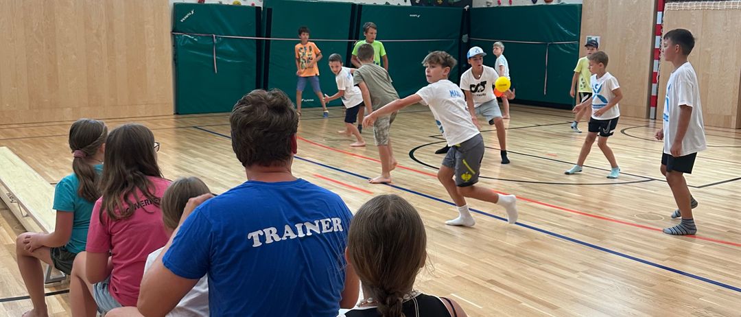 Eine Gruppe von Kindern und Erwachsenen spielt Volleyball in einer Turnhalle mit Holzboden. Einige stehen und spielen, während andere sitzen und zuschauen.