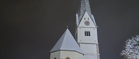 Eine hohe Kirche mit einem spitzen Turm und einer Uhr ist bei Nacht beleuchtet. Das Gebäude hat ein schneebedecktes Dach und ist von einem dunklen Himmel umgeben.