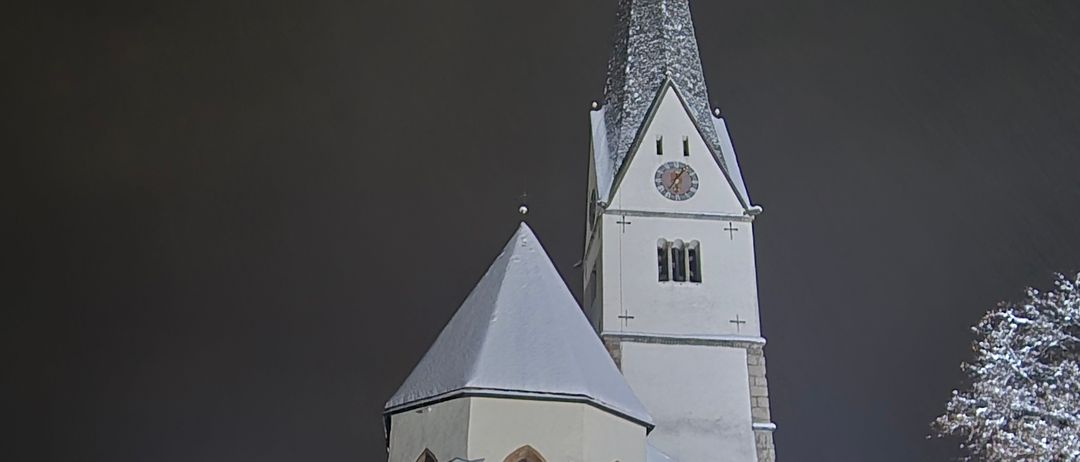 Eine hohe Kirche mit einem spitzen Turm und einer Uhr ist bei Nacht beleuchtet. Das Gebäude hat ein schneebedecktes Dach und ist von einem dunklen Himmel umgeben.