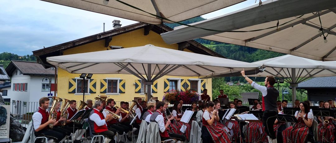 Ein Outdoor-Blasorchester spielt unter großen Schirmen. Die Musiker spielen Trompeten, Tuben und Flöten. Der Dirigent leitet von vorne.