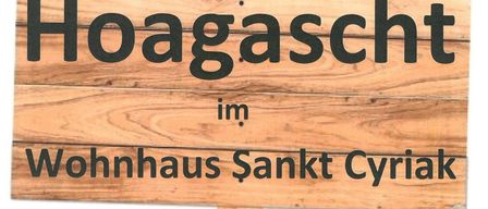 Ein Holzschild zeigt 'Hoagascht im Wohnhaus Sankt Cyriak'. Darunter steht 'Erster Mittwoch im Monat um 13:30 Uhr'. Weiter unten heißt es 'Wir freuen uns auf Ihren Besuch'.