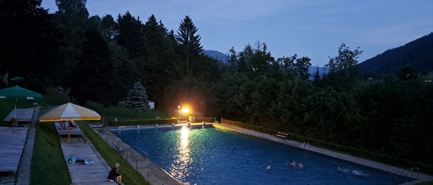 Ein Freibad mit schwimmenden Personen und anderen, die herumstehen. Bäume und Berge im Hintergrund und ein Weg, der zum Pool führt.