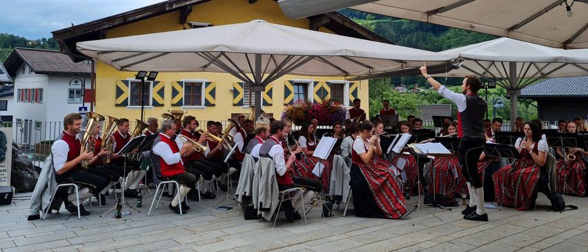 Ein Bläser- und Streicherensemble spielt im Freien, unter weißen Sonnenschirmen sitzend vor einem gelben Gebäude mit einem Berg im Hintergrund.