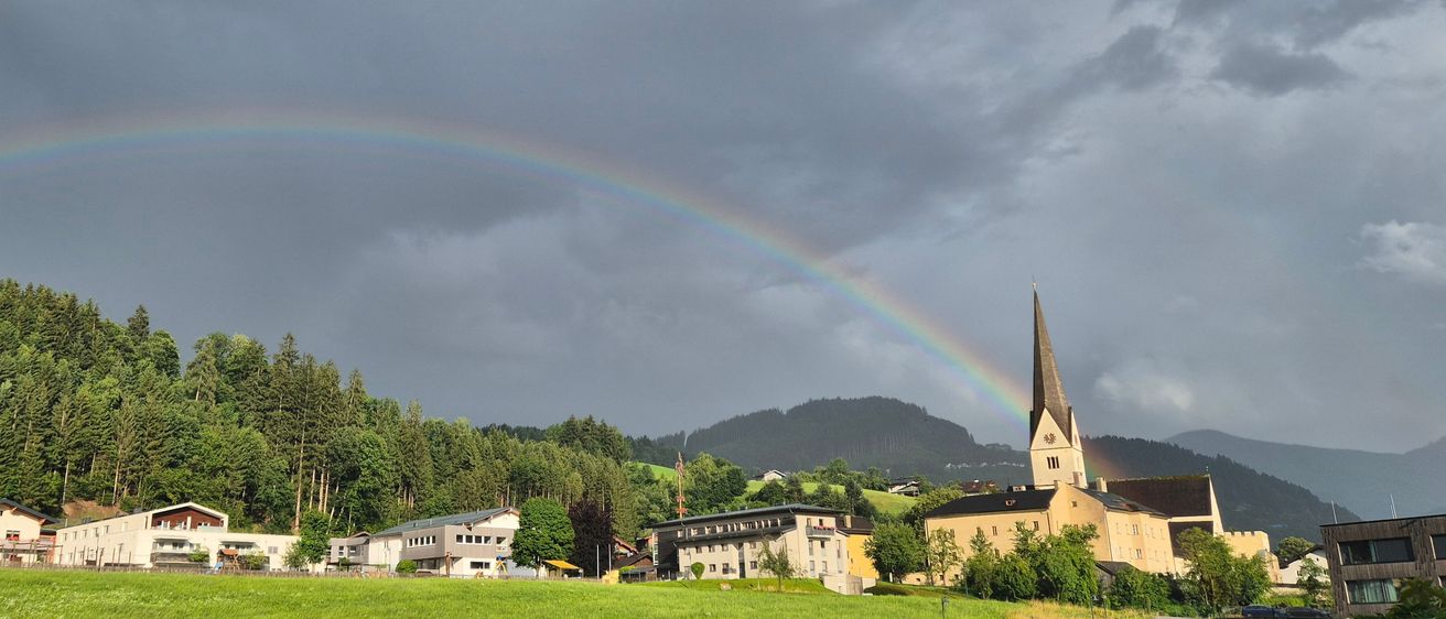 Ein leuchtender Regenbogen spannt sich über eine kleine Stadt in den Bergen, umgeben von üppigem Grün und Gebäuden.