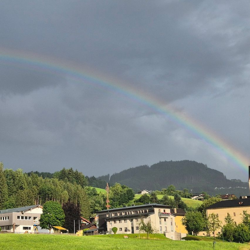 Ein leuchtender Regenbogen spannt sich über eine kleine Stadt in den Bergen, umgeben von üppigem Grün und Gebäuden.