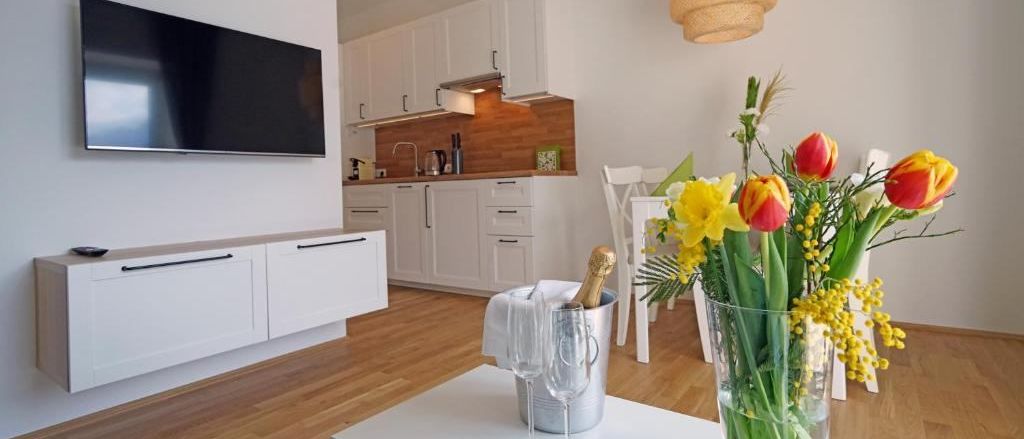 Ein modernes Wohnzimmer mit einem Flachbildfernseher an der Wand, einem Esstisch mit Blumen und einer Küche mit weißen Schränken und einer Wasserhahn.