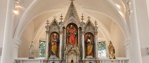 Das Innere einer Kirche zeigt einen zentralen Altar, geschmückt mit Statuen, flankiert von zwei gewölbten Fenstern. Links führt eine Tür in einen anderen Raum. Der Altar hat zwei Kerzen, und ein Kreuz ist darüber positioniert.