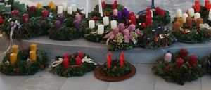 Eine Reihe von Kränzen, geschmückt mit verschiedenfarbigen Kerzen, grünen Blättern und Tannenzapfen, auf Betonstufen.