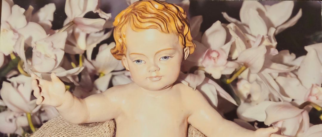 Eine Figur des Baby Jesus ruht auf einem Sacktuch, umgeben von weißen Blumen. Das Baby hat blaue Augen und einen ruhigen Ausdruck. Das Wort 'Gesegnete' ist deutlich im Bild zu sehen.
