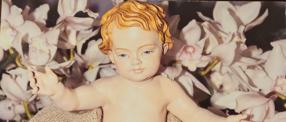 Eine Figur des Jesuskindes umgeben von weißen Blumen mit einem Jutetuch, in sitzender Position mit dem Wort 'Gesegnete' oben.