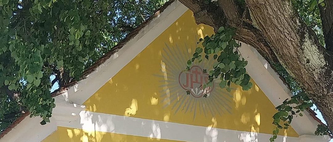 Ein gelbes Kirchengebäude mit weißen Abschlüssen und einem roten Kreuzzeichen über der Eingangstür. In der Nähe befinden sich Pflanzen und Bäume, und davor steht eine Bank.