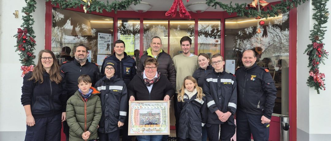 Eine Gruppe von Menschen steht vor einem Geschäft und hält einen gerahmten Gegenstand. Eine Frau in der Mitte lächelt. Hinter ihnen hat das Geschäft eine Weihnachtsdekoration.