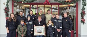 Eine Gruppe von Menschen steht vor einem Geschäft und hält einen gerahmten Gegenstand. Eine Frau in der Mitte lächelt. Hinter ihnen hat das Geschäft eine Weihnachtsdekoration.