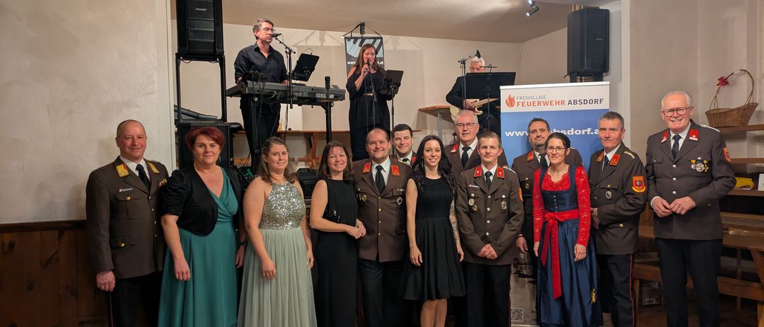 Eine Gruppe von Menschen in Uniformen und formeller Kleidung steht auf einem Holzboden vor einer Bühne mit einem Banner. Eine Band spielt im Hintergrund.
