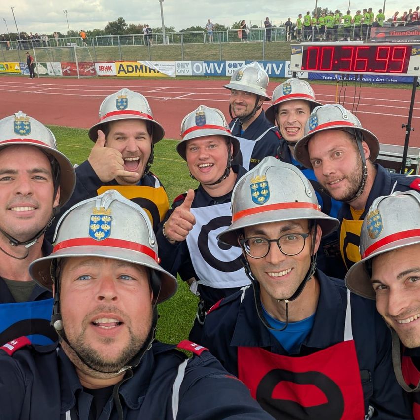 Eine Gruppe von Feuerwehrleuten in silbernen Helmen posiert für ein Foto auf einem Rasenfeld mit einer digitalen Uhr im Hintergrund.