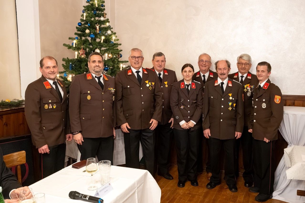 Eine Gruppe uniformierter Personen steht vor einem geschmückten Weihnachtsbaum und lächelt für ein Foto. Sie tragen Medaillen und Uniformen, mit einigen Gläsern und einem Mikrofon auf einem Tisch davor.