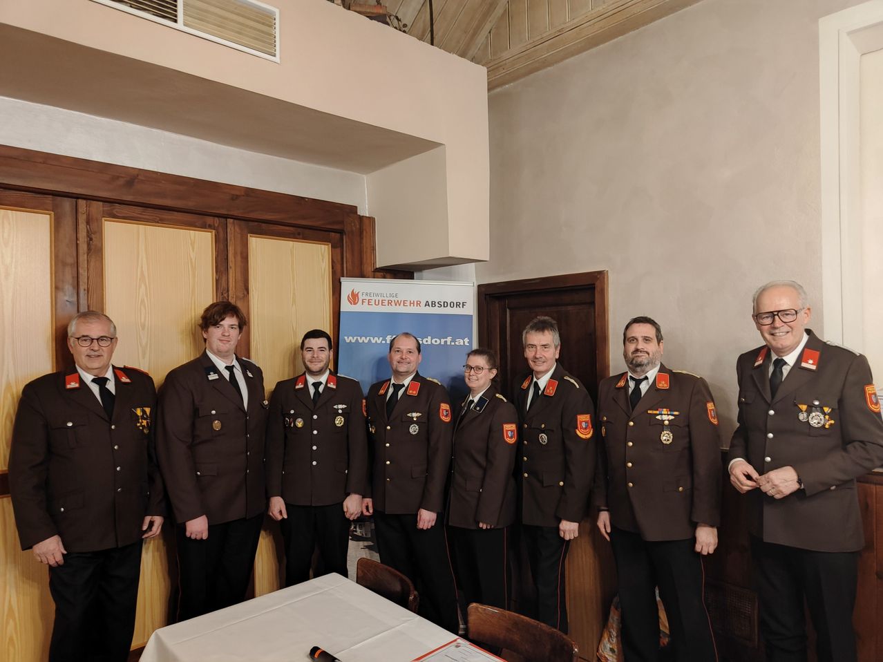 Eine Gruppe uniformierter Personen, möglicherweise Feuerwehrleute, steht zusammen vor einem Banner mit den Worten 'Feuerwehr Absdor'. Sie lächeln und posieren für ein Foto. Dahinter befindet sich eine Holztür mit einem Glasfenster.