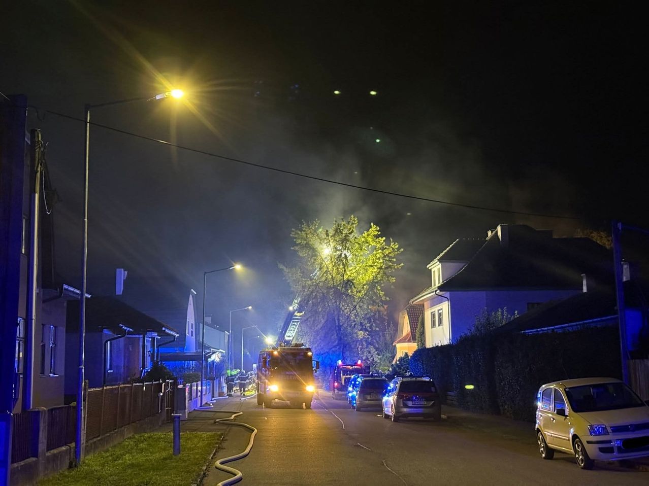 Einsatzfahrzeuge, einschließlich eines Feuerwehrwagens, sind auf der Straße geparkt. Ein Feuer brennt in der Nähe der Häuser, wobei Rauch in den Himmel steigt.