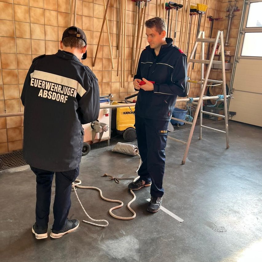 Zwei Männer in Uniformen stehen in einer Garage, einer hält ein rotes Objekt, der andere steht ihm gegenüber. Hinter ihnen befinden sich Werkzeuge und Ausrüstung an den Wänden und am Boden.