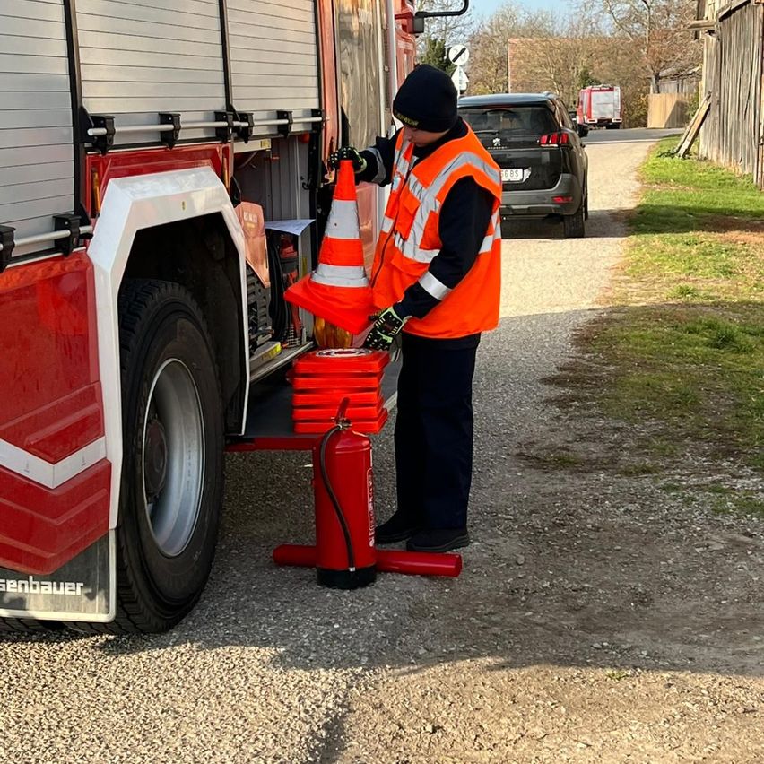 Ein Feuerwehrmann steht neben einem Feuerwehrauto, hält einen Verkehrskegel und einen Feuerlöscher. Das Auto ist auf einer Kiesstraße geparkt, mit einem Auto und einem anderen Lastwagen im Hintergrund.
