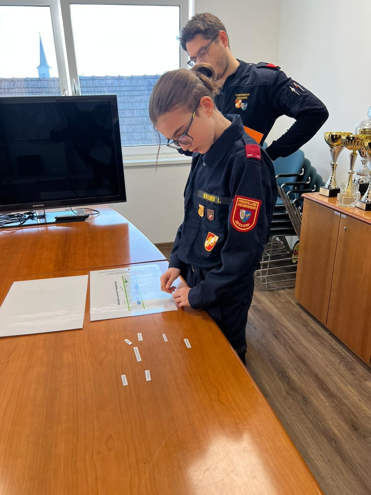 Eine Frau in Uniform liest ein Dokument an einem Tisch, während eine andere Person in Uniform hinter ihr steht. Trophäen stehen auf einem nahen Regal.