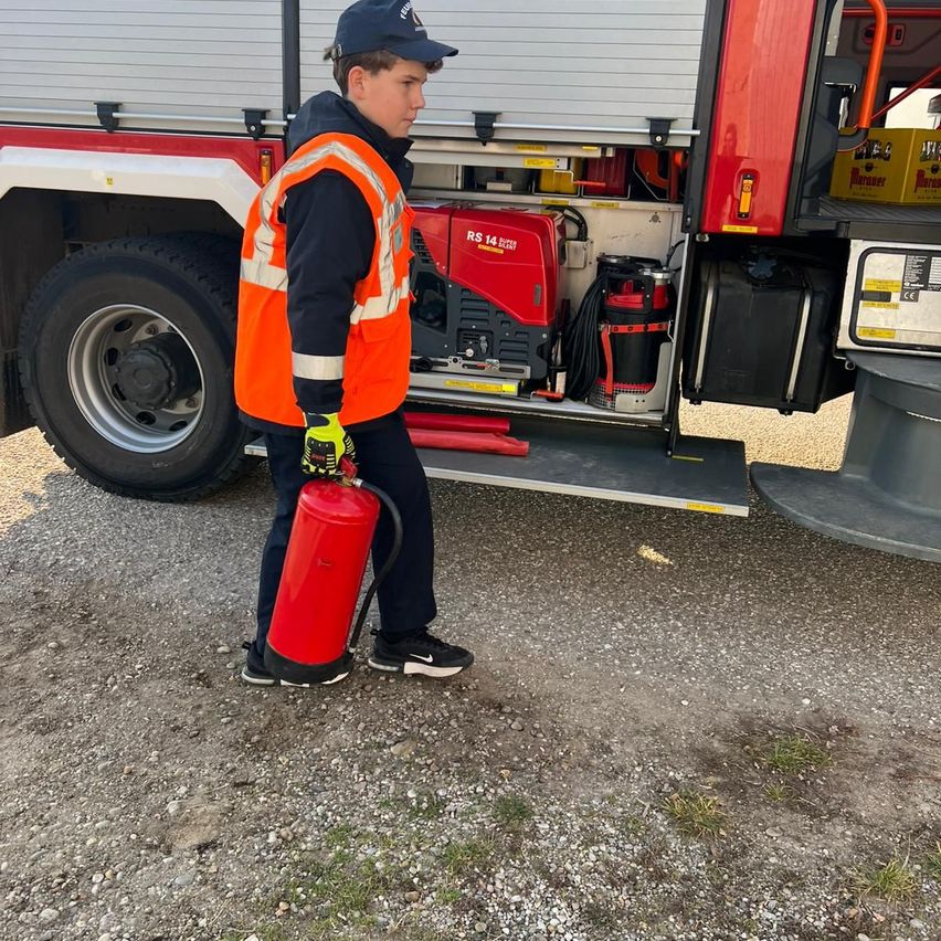 Ein junger Junge in einer Warnweste hält einen roten Feuerlöscher vor einem Feuerwehrwagen.