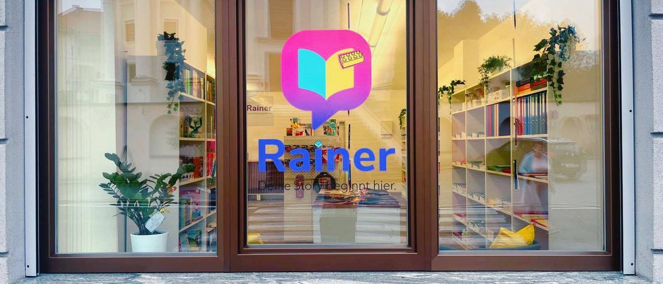 Schaufenster eines Buchladens mit dem Namen 'Rainer' in Pink. Regale voller Bücher, eine Topfpflanze und eine Frau im Hintergrund. Die Fenster spiegeln das Innere und Äußere wider.