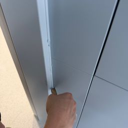 Eine Hand hält ein Werkzeug, möglicherweise ein Schaber, gegen eine Wandplatte. Die Platte hat eine hellblaue Farbe und einen leicht gebogenen Rand.