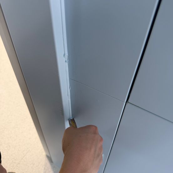 Eine Hand hält ein Werkzeug, möglicherweise ein Schaber, gegen eine Wandplatte. Die Platte hat eine hellblaue Farbe und einen leicht gebogenen Rand.