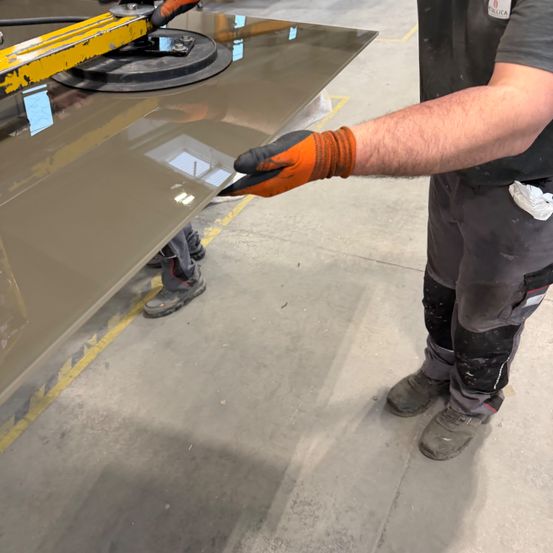 Ein Arbeiter in einer Fabrik hält eine Glasplatte mit einem Spannwerkzeug, trägt orangefarbene Handschuhe und Schutzausrüstung.