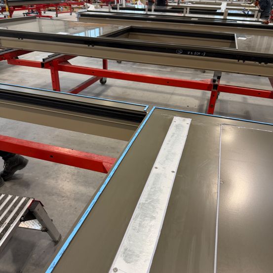 Ein industrielles Umfeld mit roten und silbernen Metallrahmen, die Glasplatten halten. Eine Person steht neben einer Leiter. Der Boden ist aus Beton.