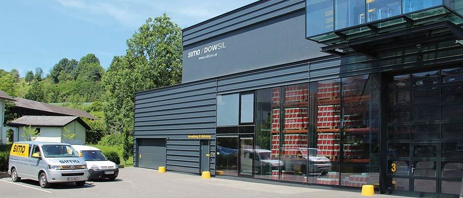 Ein modernes Industriegebäude mit Glaswänden und einem Schild, das 'SIMO/DOWSIL' steht neben einer Straße mit geparkten Fahrzeugen, umgeben von Grün.
