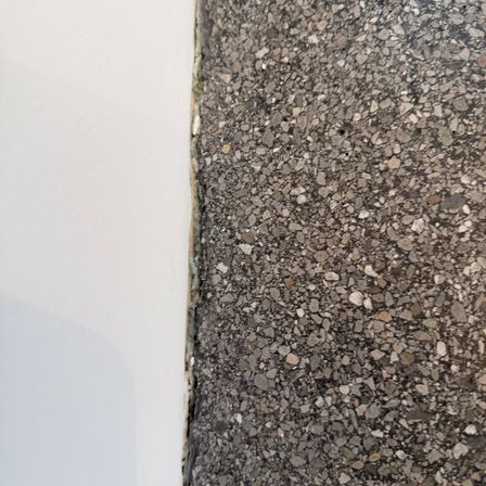 Nahaufnahme eines strukturierten Betonbodens mit sichtbaren Zuschlagstoffen. Der Boden trifft auf eine weiße Wand links.