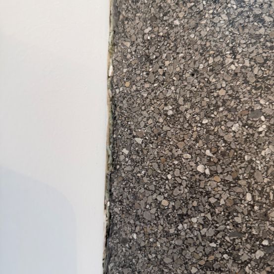 Nahaufnahme eines strukturierten Betonbodens mit sichtbaren Zuschlagstoffen. Der Boden trifft auf eine weiße Wand links.