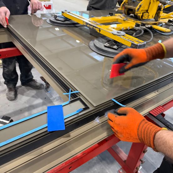 Zwei Personen in orangefarbenen Handschuhen arbeiten an einer Metallplatte, eine hält ein rotes Objekt und die andere ein blaues Objekt. Die Metallplatte befindet sich auf einer gelben Maschine mit rotem Stützrahmen.