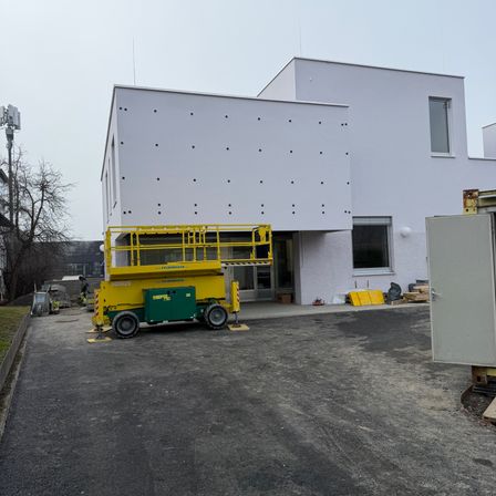 Ein weißes Gebäude mit einem gelben Scherenarbeitsbühne vor ihm. Es gibt Baumaterialien und einen Baum in der Nähe.