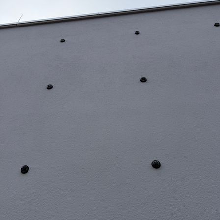 Eine graue Wand mit mehreren schwarzen kreisförmigen Objekten gleichmäßig auf ihrer Oberfläche verteilt. Der Himmel ist klar und deutet auf einen sonnigen Tag hin.