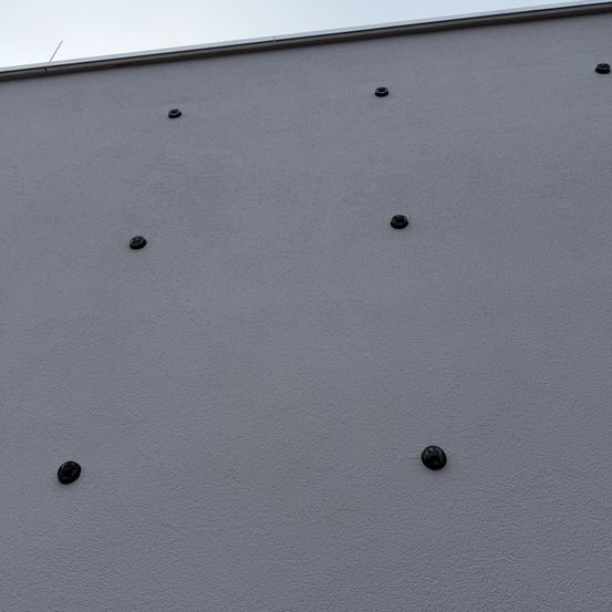 Eine graue Wand mit mehreren schwarzen kreisförmigen Objekten gleichmäßig auf ihrer Oberfläche verteilt. Der Himmel ist klar und deutet auf einen sonnigen Tag hin.