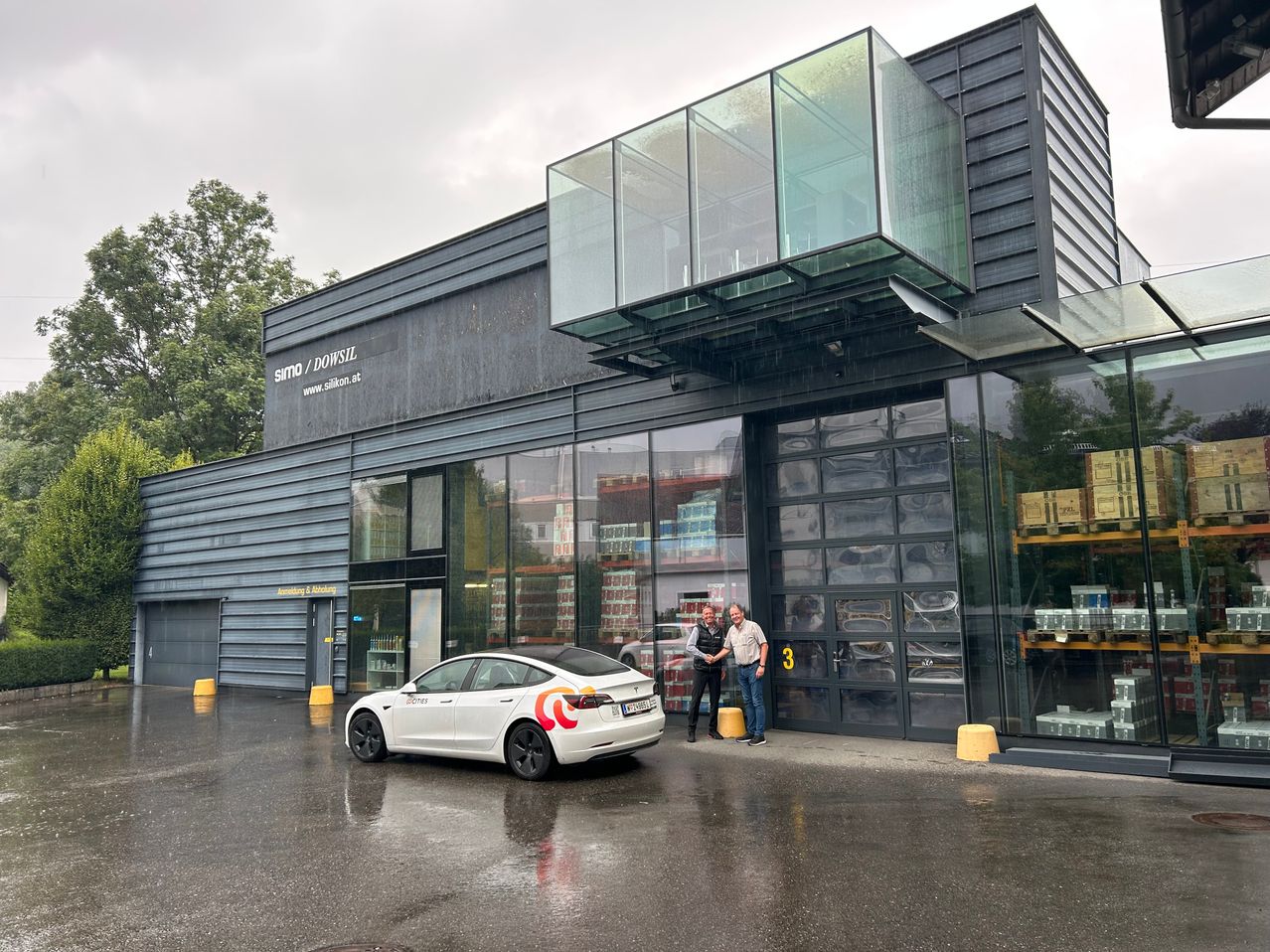 Zwei Männer stehen vor einem modernen Gebäude mit Glasfassade, in der Nähe eines Tesla Model 3. Das Gebäude trägt ein Schild mit der Aufschrift 'simo / DOWSIL'. Durch die Glasfenster sind Regale und Produkte zu sehen.