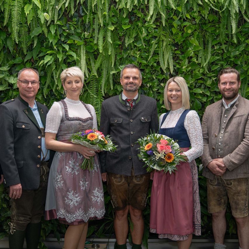 Fünf Personen posieren für ein Foto vor einer grünen Wand mit Pflanzen. Sie tragen alle traditionelle deutsche Kleidung. Die beiden Frauen halten Blumenstrauße.