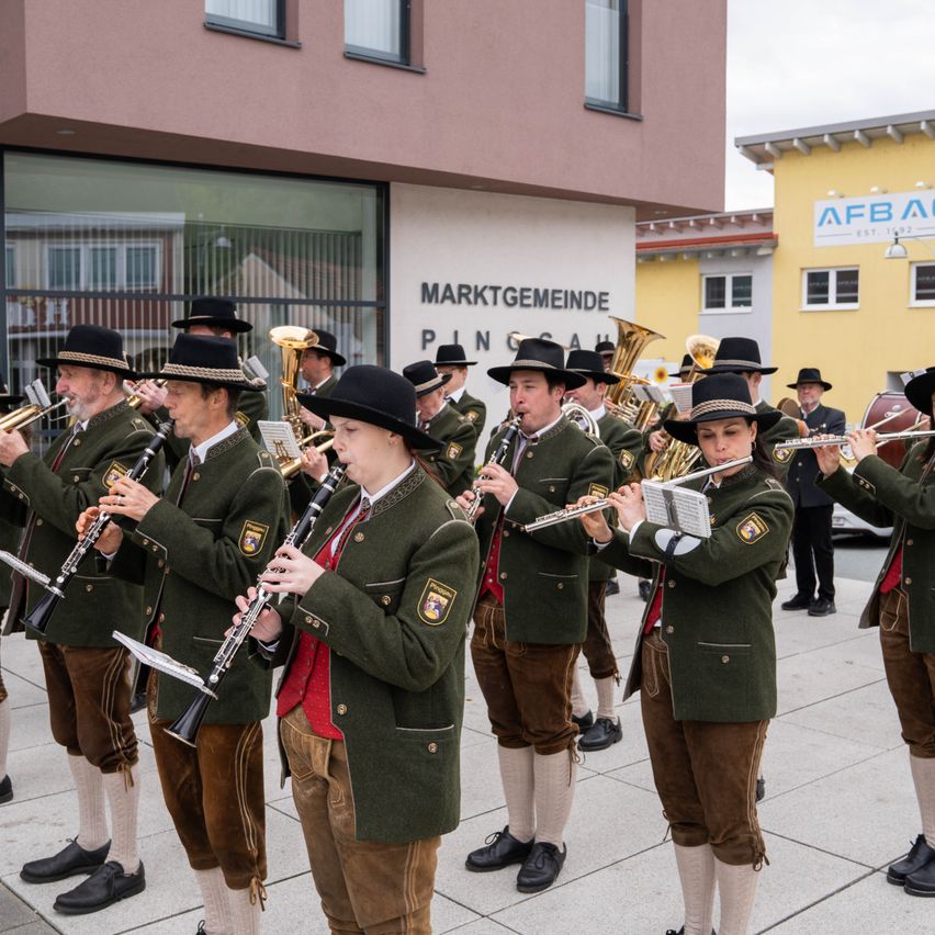 Eine Gruppe von Musikern in grünen Uniformen spielt ihre Instrumente vor einem Gebäude mit der Aufschrift 'Marktgemeinde Pinggau'.